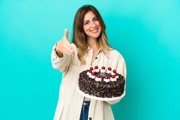 Donna caucasica che tiene la torta di compleanno isolata su fondo blu che stringe la mano per chiudere un buon affare