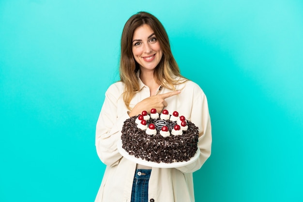 Donna caucasica che tiene la torta di compleanno isolata su sfondo blu che punta al lato per presentare un prodotto