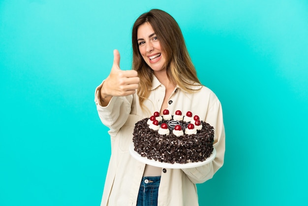 Donna caucasica che tiene la torta di compleanno isolata su sfondo blu con il pollice in alto perché è successo qualcosa di buono