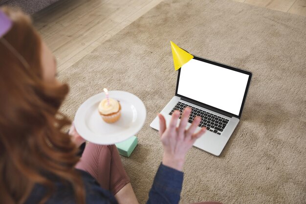Donna caucasica che trascorre del tempo a casa, in soggiorno, utilizzando il laptop, festeggiando e tenendo in mano un cupcake con una candela. Distanziamento sociale durante il blocco della quarantena del coronavirus Covid 19.