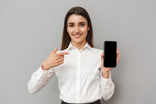 Donna caucasica con lunghi capelli castani facendo pubblicità e presentando lo schermo copyspace nero del telefono cellulare, isolato sopra il muro grigio