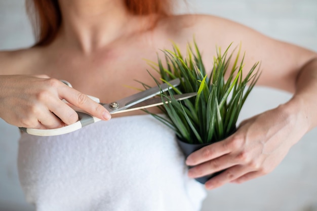 Donna caucasica dai capelli rossi senza volto avvolta in un asciugamano di spugna bianco taglia una pianta in vaso con le forbici Concetto di rimozione dei peli delle ascelle Procedura di autodepilazione Tagliare la vegetazione in eccesso