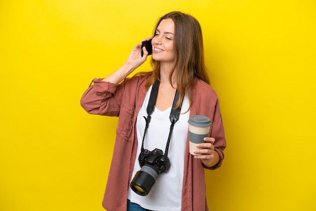 Donna caucasica del giovane fotografo isolata su fondo giallo che tiene il caffè da portare via e un cellulare