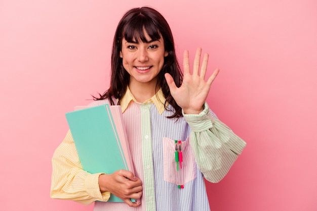 Donna caucasica del giovane studente che tiene i libri isolati sulla parete rosa sorridente allegro che mostra il numero cinque con le dita.