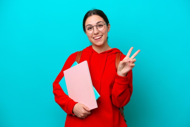 Donna caucasica del giovane studente isolata su fondo blu che sorride e che mostra il segno di vittoria