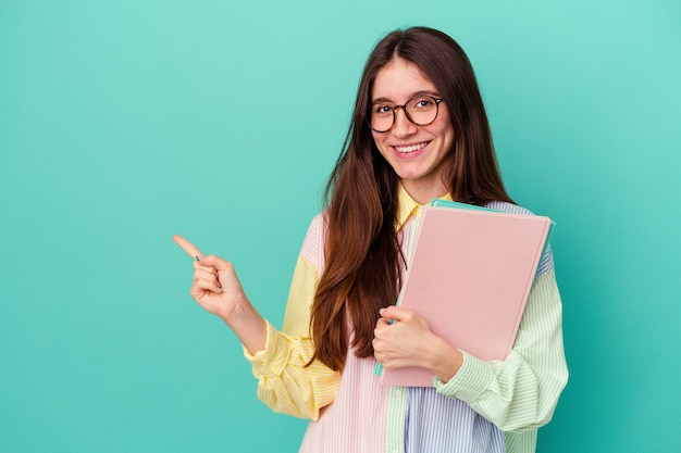 Donna caucasica del giovane studente isolata sulla parete blu che sorride e che indica da parte, mostrando qualcosa nello spazio vuoto.