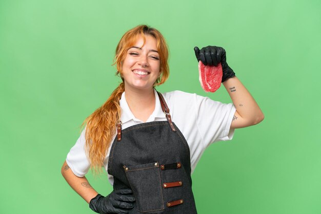 Donna caucasica del macellaio che indossa un grembiule e serve carne tagliata fresca isolata su sfondo chroma key dello schermo verde in posa con le braccia all'anca e sorridente