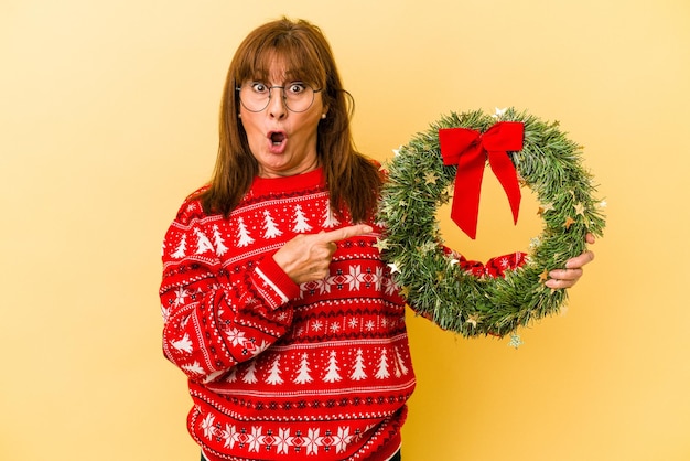 Donna caucasica di mezza età che celebra il Natale tenendo la corona isolata su sfondo giallo che punta al lato