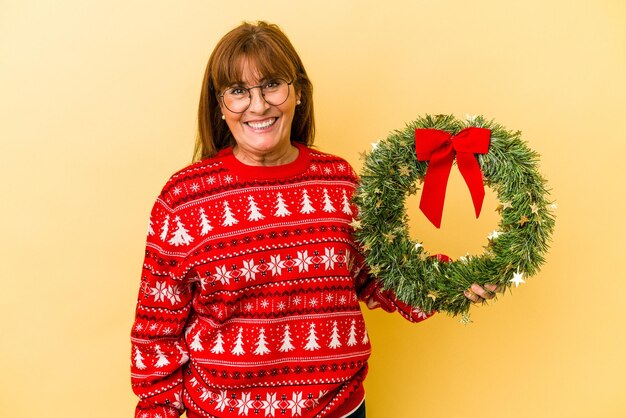 Donna caucasica di mezza età che celebra il Natale tenendo la corona isolata su sfondo giallo felice, sorridente e allegro.