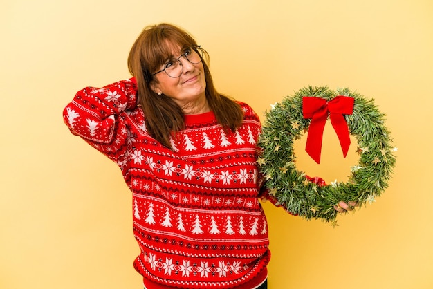 Donna caucasica di mezza età che celebra il Natale tenendo la corona isolata su sfondo giallo toccando la parte posteriore della testa, pensando e facendo una scelta.