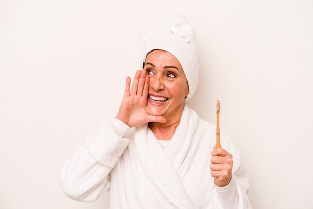 Donna caucasica di mezza età che indossa un accappatoio con spazzolino da denti isolato su sfondo bianco che grida e tiene il palmo vicino alla bocca aperta