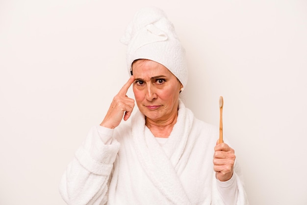 Donna caucasica di mezza età che indossa un accappatoio con spazzolino da denti isolato su sfondo bianco che punta il tempio con il pensiero del dito concentrato su un compito