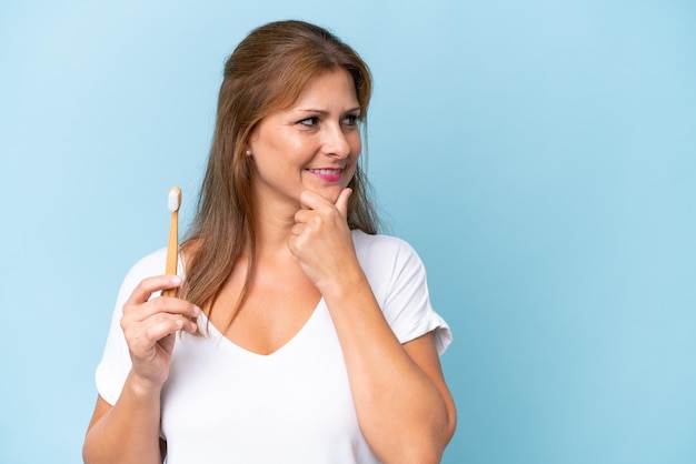 Donna caucasica di mezza età che lava i denti isolati su sfondo blu pensando un'idea e guardando il lato