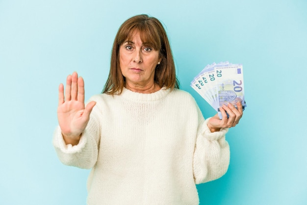 Donna caucasica di mezza età che tiene banconote isolate su sfondo blu in piedi con la mano tesa che mostra il segnale di stop, impedendoti.