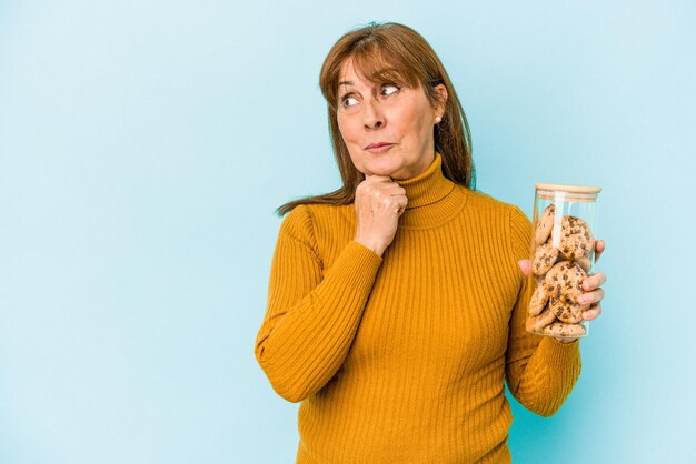 Donna caucasica di mezza età che tiene il barattolo di biscotti isolato su sfondo blu guardando lateralmente con espressione dubbiosa e scettica.