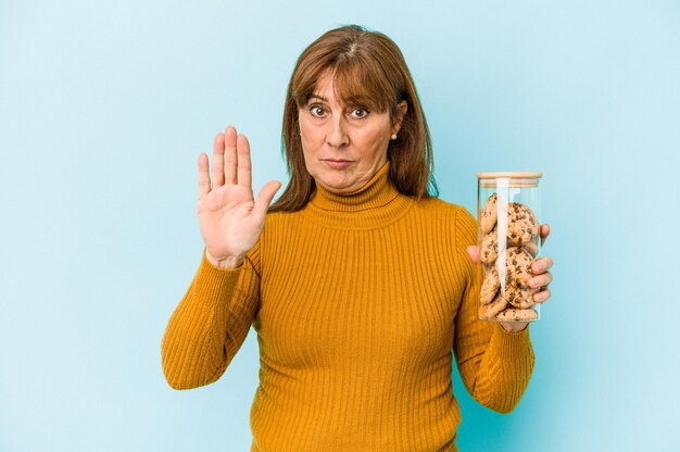 Donna caucasica di mezza età che tiene il barattolo di biscotti isolato su sfondo blu in piedi con la mano tesa che mostra il segnale di stop, impedendoti.