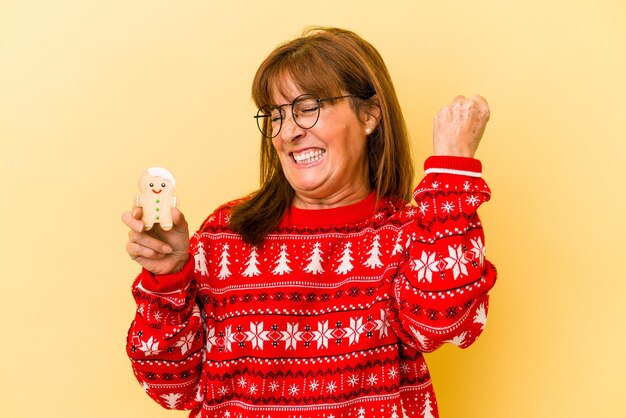 Donna caucasica di mezza età che tiene in mano un cookie di Natale isolato su sfondo giallo alzando il pugno dopo una vittoria, concetto vincitore.