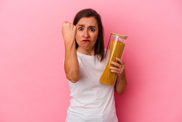 Donna caucasica di mezza età che tiene un vasetto di pasta isolato su sfondo rosa che mostra il pugno alla telecamera, espressione facciale aggressiva.