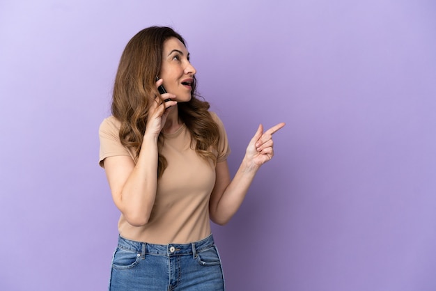 Donna caucasica di mezza età che utilizza il telefono cellulare isolato su sfondo viola puntando il dito di lato e presentando un prodotto