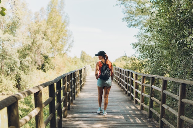 Donna caucasica di mezza età in abiti estivi che attraversa un ponte su un sentiero rurale.