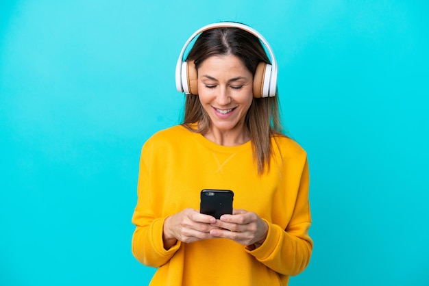 Donna caucasica di mezza età isolata su sfondo blu ascoltando musica e guardando al cellulare