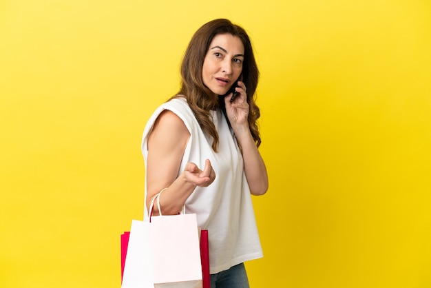Donna caucasica di mezza età isolata su sfondo giallo tenendo le borse della spesa e chiamando un amico con il suo cellulare