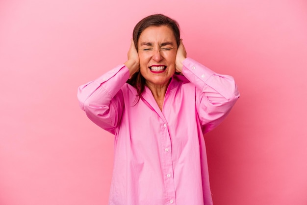 Donna caucasica di mezza età isolata su sfondo rosa che copre le orecchie con le mani cercando di non sentire un suono troppo forte