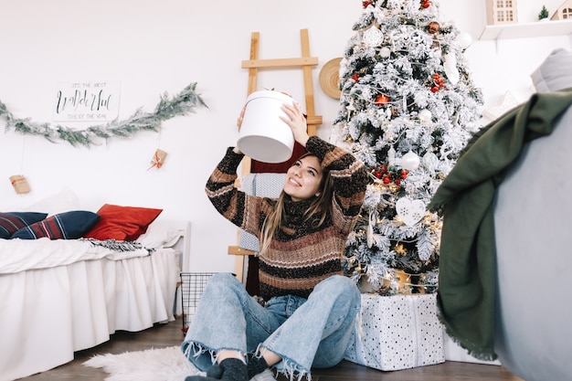 Donna caucasica felice che tiene grande regalo di Natale mentre era seduto sul pavimento a casa vicino all'albero di Natale.