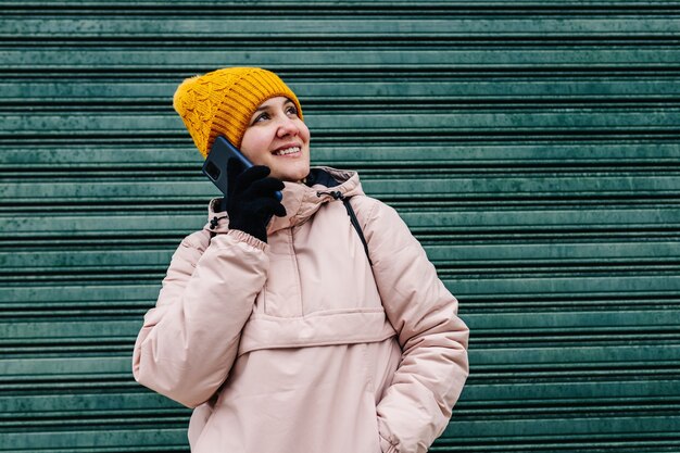 Donna caucasica in abiti invernali, parlando al cellulare accanto al muro verde.