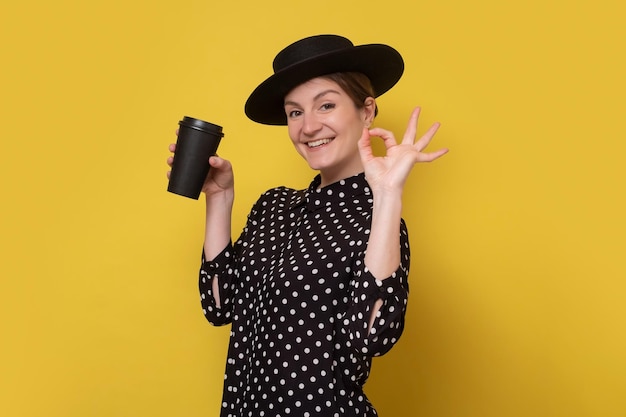 Donna caucasica in cappello nero che tiene una tazza di caffè che mostra un gesto ok isolato sulla parete gialla Studio girato