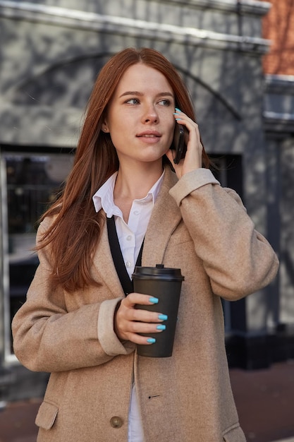 Donna caucasica in elegante cappotto beige che chiama dallo smartphone bevendo caffè