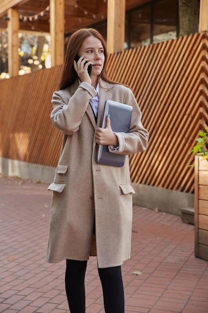 Donna caucasica in elegante cappotto beige che chiama dallo smartphone che tiene il laptop