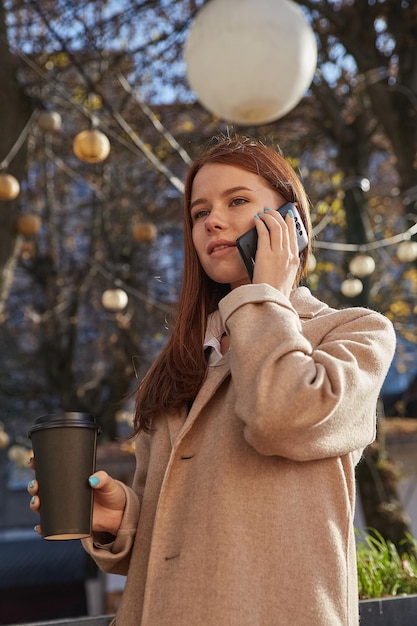 Donna caucasica in elegante cappotto beige che chiama tramite smartphone, bevendo caffè