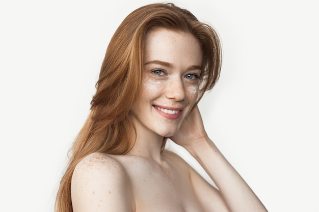 Donna caucasica lentigginosa con capelli rossi che sorride alla macchina fotografica, in posa con spalle nude e crema sotto gli occhi