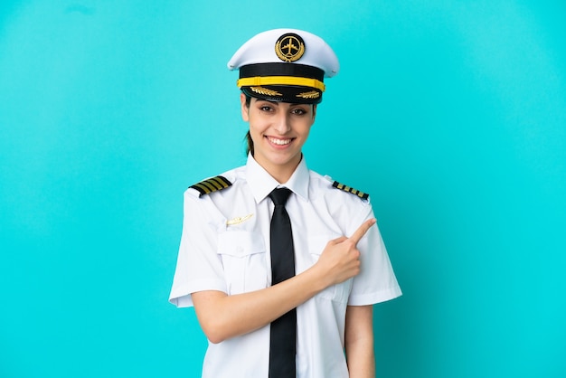 Donna caucasica pilota di aeroplano isolata su sfondo blu che punta al lato per presentare un prodotto