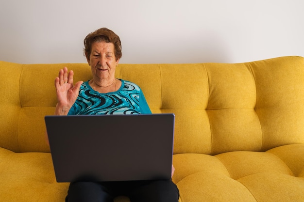Donna caucasica più anziana che effettua una videochiamata seduta su un divano giallo Indossa una maglietta blu