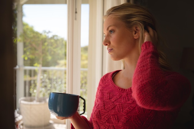 Donna caucasica premurosa in piedi vicino alla finestra che tiene la tazza di caffè che guarda al giardino soleggiato. passare il tempo libero in casa.