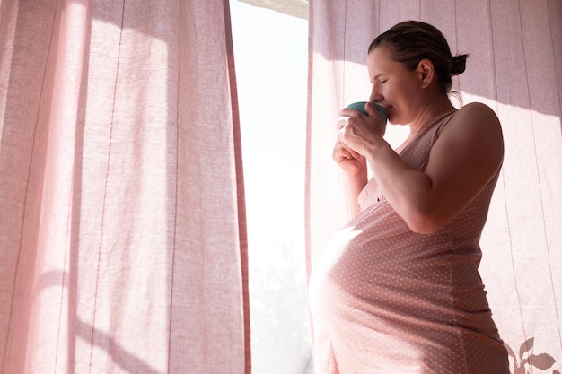 Donna caucasica rilassante bere tè caldo in piedi vicino alla finestra