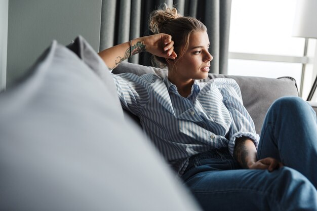 donna caucasica seria in camicia a righe che guarda da parte mentre è seduta sul divano in soggiorno