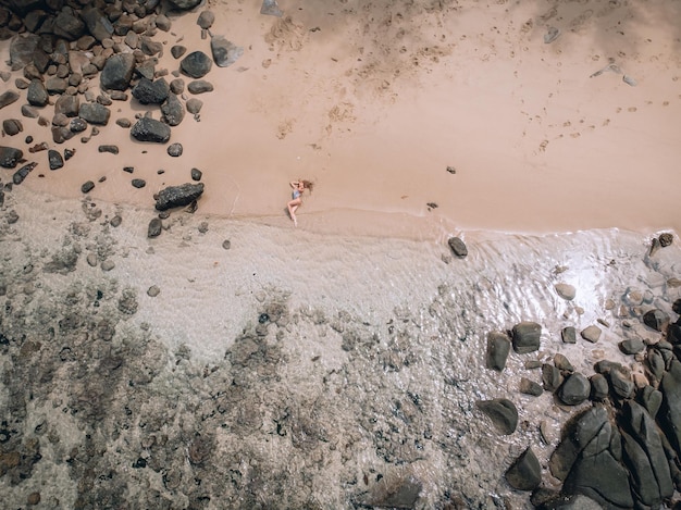 Donna caucasica sexy sulla spiaggia sulla sabbia vicino alle onde e alle pietre. La bella donna si copre i seni nudi con le mani. Phuket. Tailandia. Vista aerea dall'alto.