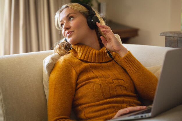 Donna caucasica sorridente che si rilassa sul divano del soggiorno indossando le cuffie e utilizzando il computer portatile. trascorrere il tempo libero a casa con la tecnologia.