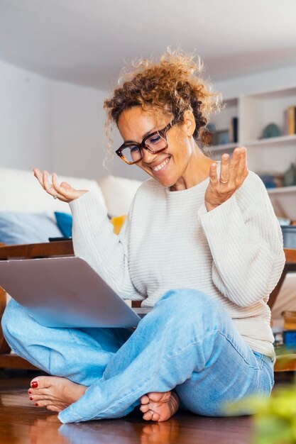 Donna caucasica sorridente che usa il portatile per una videochiama mentre è seduta a casa sul pavimento Femmina felice che ha una riunione online con un cliente o parla con amici o familiari Spazio di copia Comunicazione