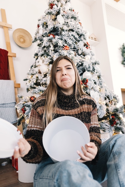 Donna caucasica triste che tiene grande regalo di Natale vuoto mentre era seduto sul pavimento a casa vicino all'albero di Natale.