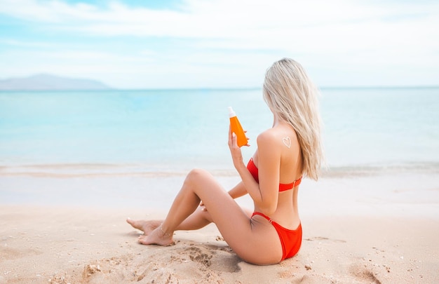 Donna che applica crema solare sul corpo abbronzato In forma cuore amore per proteggere la sua pelle Ragazza che usa crema solare Donna che tiene crema solare idratante crema solare in una giornata di sole con mare sfocato sullo sfondo