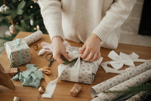 Donna che avvolge un elegante regalo di Natale con nastro e ramo di abete su un tavolo di legno con decorazioni festive in una stanza scandinava decorata in modo moderno Buon Natale Mani che impacchettano il regalo da vicino