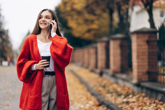 Donna che beve caffè e usa il telefono e cammina in strada d'autunno