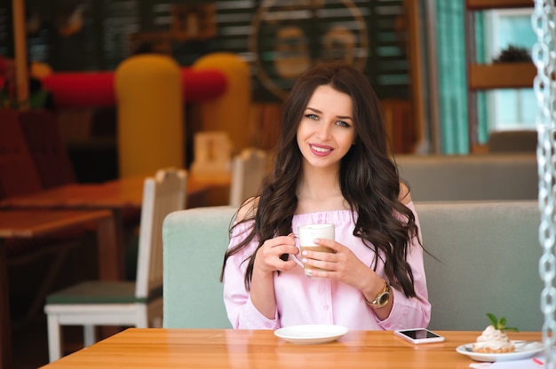 Donna che beve il caffè caldo del cappuccino e che mangia dolce in un caffè.