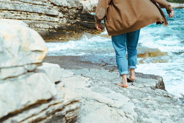 Donna che cammina dalla spiaggia rocciosa del mare al concetto spensierato di vacanza estiva soleggiata giornata ventosa onde sullo sfondo