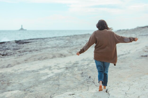 Donna che cammina dalla spiaggia rocciosa del mare nel faro dei jeans bagnati sul concetto spensierato delle vacanze estive del tempo ventoso del fondo