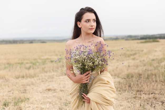 Donna che cammina nel campo di erba secca dorata Bellezza naturale del ritratto Bella ragazza in un campo di grano Giovane donna in un vestito beige che tiene un mazzo di fiori di campo